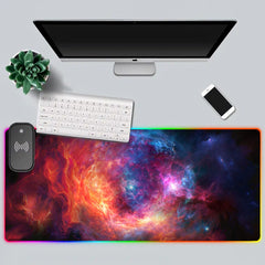 Top-down white desk setup with The Cradle RGB deskmat, keyboard, monitor, phone, and plant.